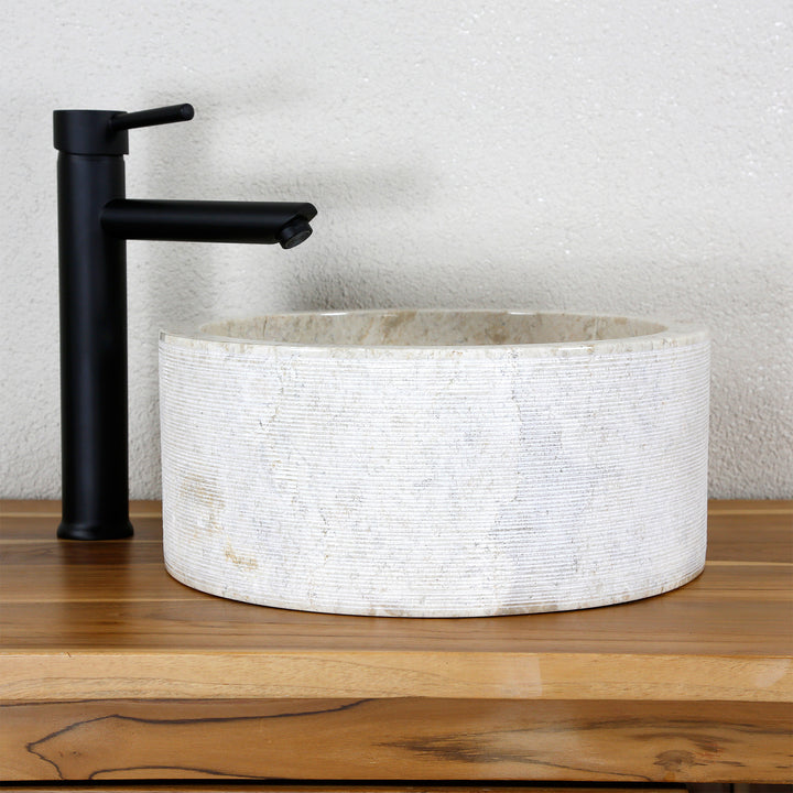 Lavabo à poser rond en pierre naturelle claire avec une structure aux lignes fines, associé à un robinet autonome noir.