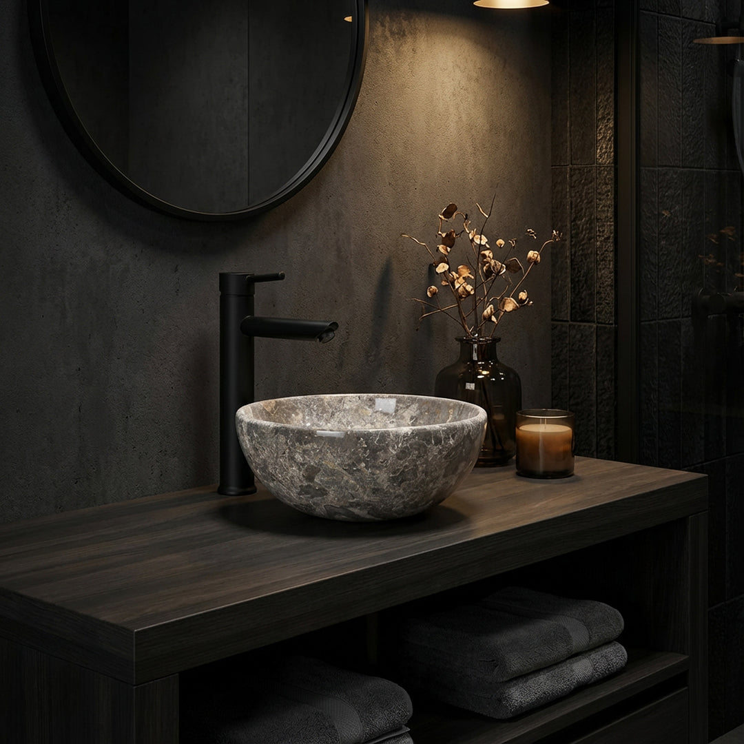 Vasque à poser ronde en pierre naturelle au grain gris-brun sur une console en bois sombre dans la salle de bains éclairée d'ambiance, associée à des robinetteries noires, un miroir et des accessoires subtils