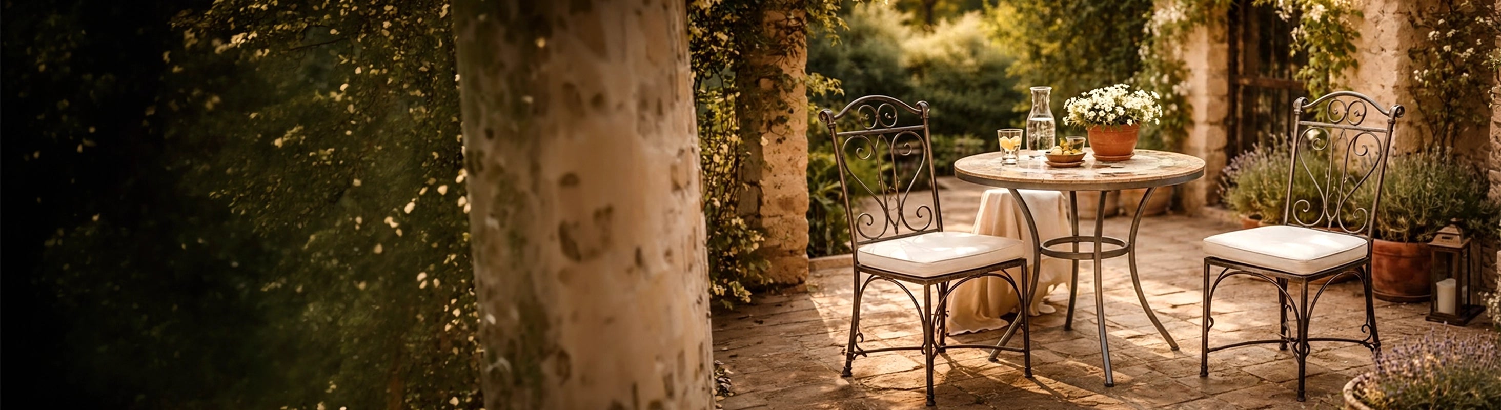 Chaises en fer – Schmiedeeiserne Stühle mit Sitzkissen an rundem Tisch auf mediterraner Terrasse mit Natursteinboden und Pflanzen