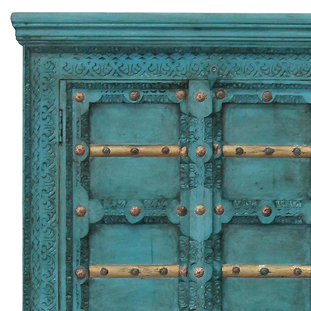Gros plan des ornements sculptés turquoise, des traverses dorées et des ferrures sur les portes de l’armoire.