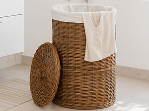 Haut panier à linge en rotin brun avec sac clair et linge beige dans la salle de bains, couvercle posé devant
