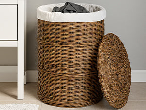 Panier à linge en rotin brun avec bord en tissu clair et vêtements foncés près d’une armoire blanche, couvercle à droite