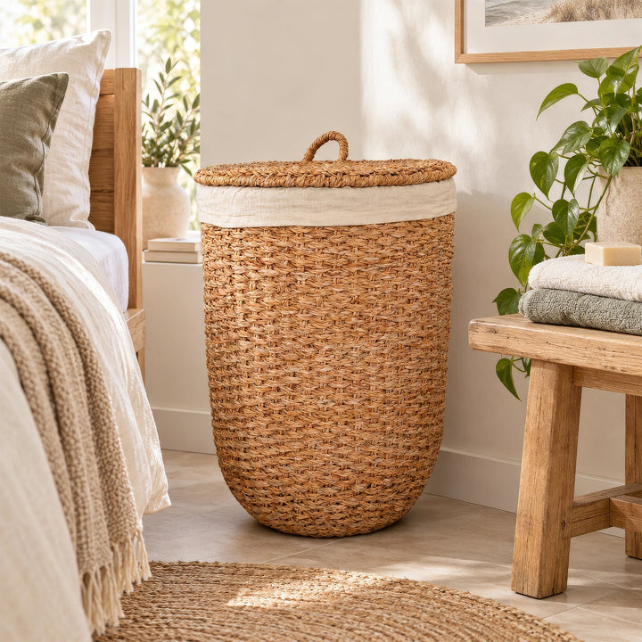 Panier à linge avec couvercle, en jonc de mer de couleur naturelle et doté d’un empiècement en tissu clair, placé entre le lit et le banc en bois dans la chambre.