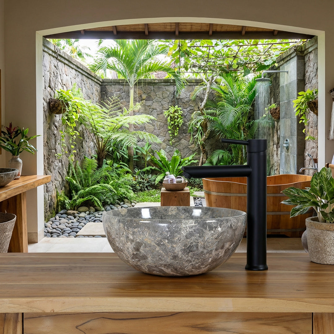 Pierre naturelle ronde Lavabo avec des grains gris-brun sur un lavabo en bois dans la salle de bain spa ouverte avec vue sur une cour verdoyante, combiné avec un robinet autonome noir, un mur en pierre naturelle et une baignoire en bois